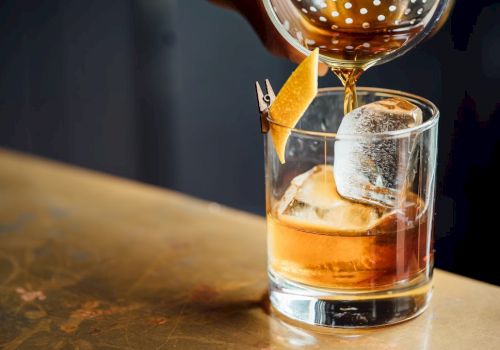 A glass tumbler with whiskey on the rocks, a large ice cube, and an orange peel garnish being twisted, with a drink being poured from a carafe.