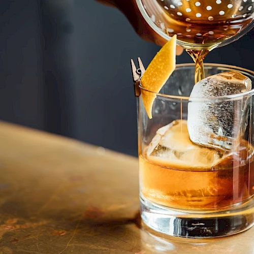 A glass tumbler with whiskey on the rocks, a large ice cube, and an orange peel garnish being twisted, with a drink being poured from a carafe.