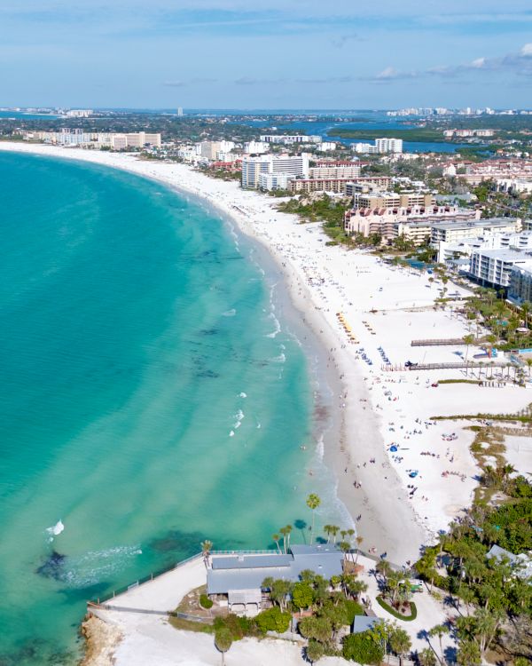 Aerial view of a curved white-sand beach along a turquoise sea, with resort hotels and palm trees lining the coast and clear blue skies above.