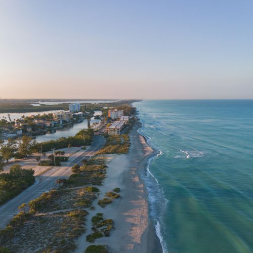 Coastal view of a shoreline with turquoise sea, gentle waves, and a few buildings inland, under a clear blue sky.