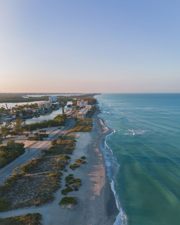 Coastal view of a shoreline with turquoise sea, gentle waves, and a few buildings inland, under a clear blue sky.