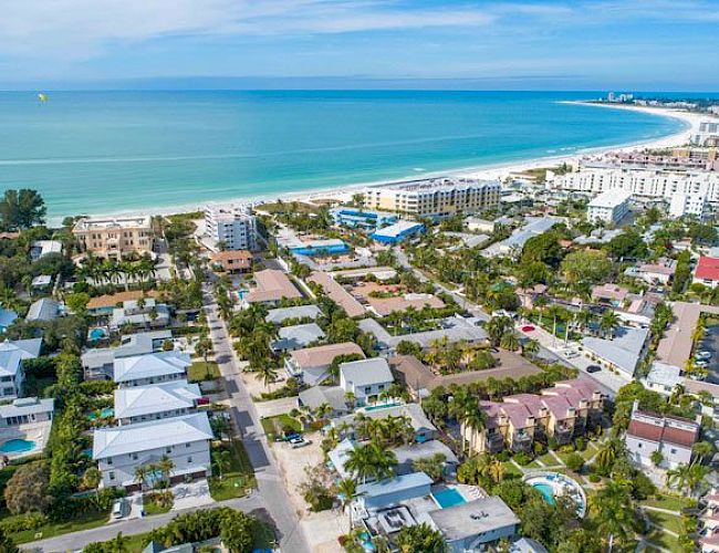 Aerial view of a coastal town with palm trees, row houses, and low-rise buildings along a curved white-sand beach and turquoise sea, sunny and inviting.