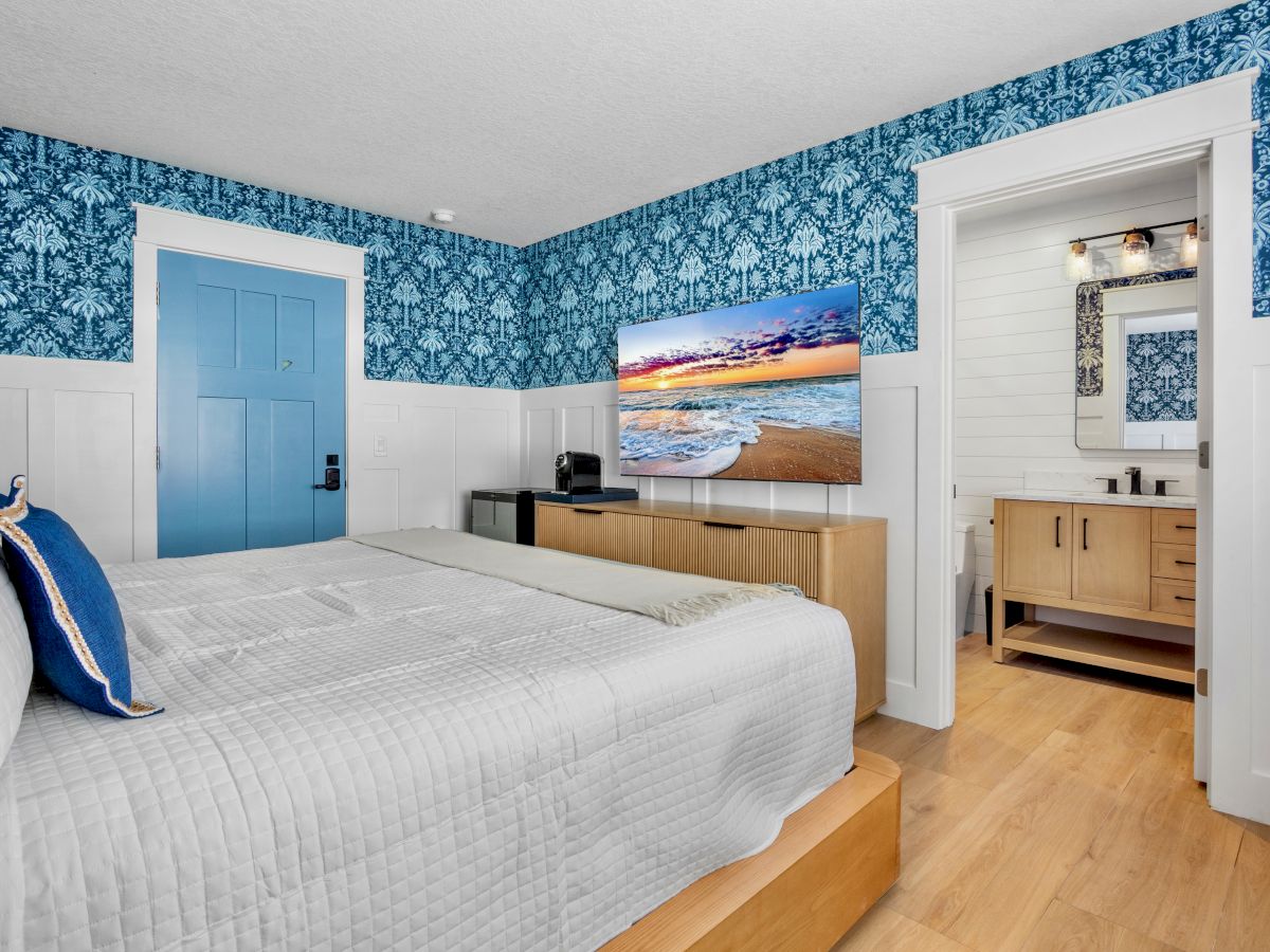 A bright hotel-style bedroom with blue patterned wallpaper, a white bed, blue door, wooden furniture, and a small seating area by a dresser, ending with a sunset art piece.