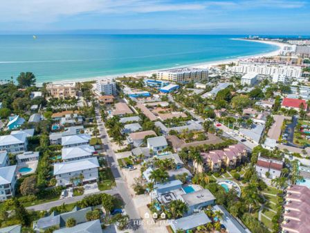Aerial view of a coastal neighborhood with white buildings, palm trees, and a clear blue ocean along the shore. The town stretches between the beach and inland streets.