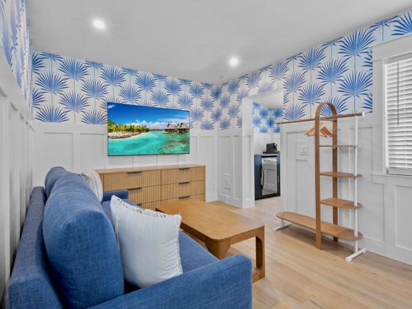 A small living room with blue patterned wallpaper, white paneling, a blue sofa, a wooden coffee table, a TV on a dresser, and a wooden coat rack.
