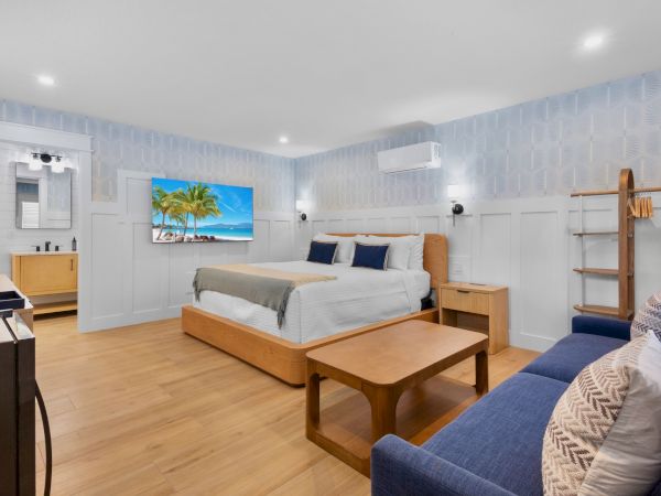 A bright hotel-style bedroom with a king bed, blue accents, a sofa, coffee table, flat TV on the wall, desk, and air conditioner&mdash;cozy and modern.