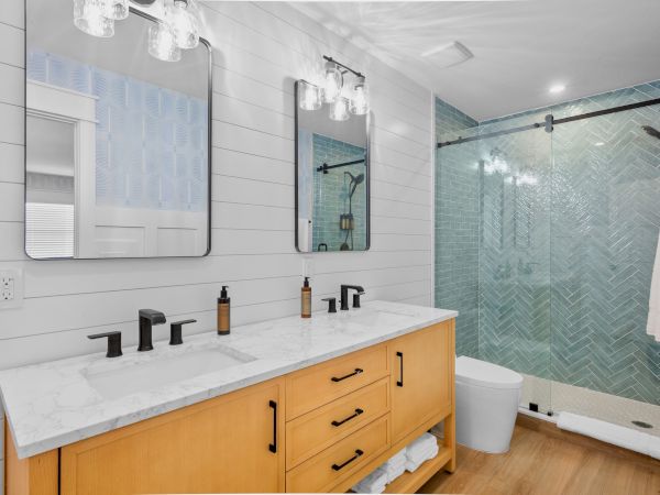 A modern bathroom with a double vanity, black faucets, a light wood cabinet, white marble countertop, and a glass shower with chevron tiles.