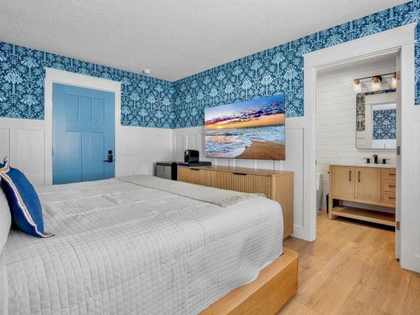 A bright hotel room with blue patterned wallpaper, a white bed, blue accents, a door, a wooden dresser, a large TV, and an adjacent bathroom.