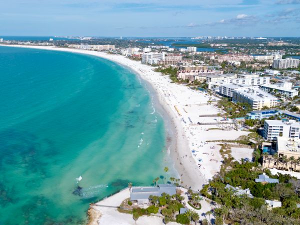 A sunny coastal scene: turquoise sea, white sandy beach, and a row of mid-rise hotels along the shoreline, with clear blue skies above.