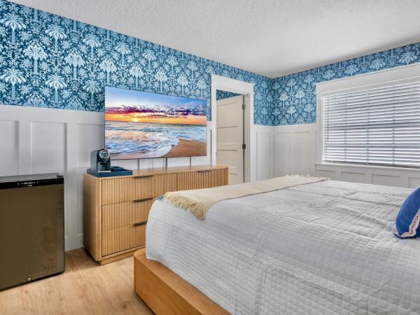 A cozy bedroom with blue floral wallpaper, white wainscoting, a wooden dresser, a TV on top, a small fridge on the left, a bed with white bedding and blue pillows, and a window with blinds.