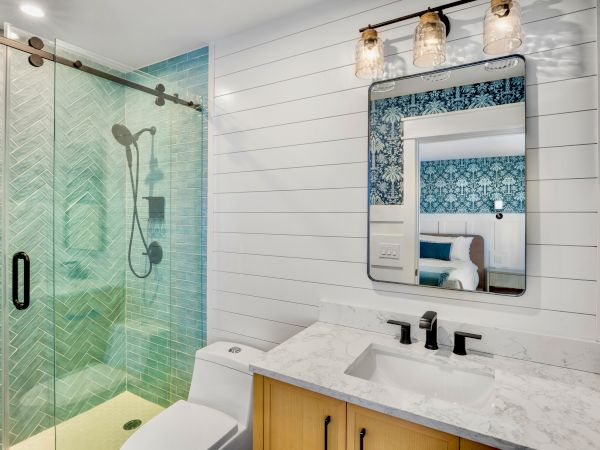 A modern bathroom with a glass shower, white shiplap walls, a marble countertop, wooden vanity, and a decorative framed mirror above dual sinks.