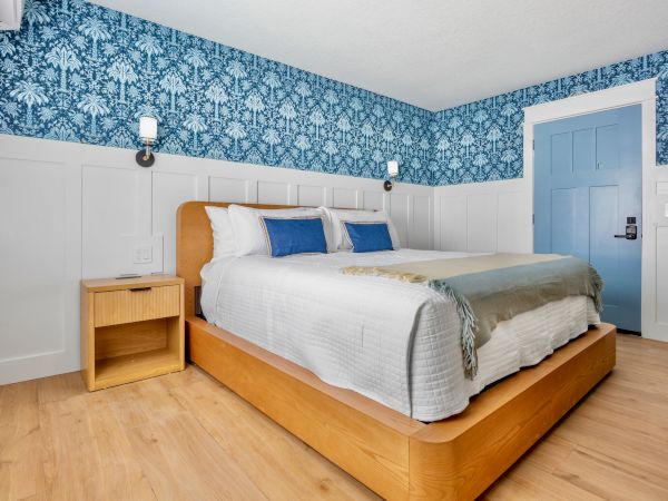 A bright hotel-style bedroom with a wooden platform bed, white bedding, blue throw pillows, blue patterned wallpaper, light wood floors, and matching nightstands.