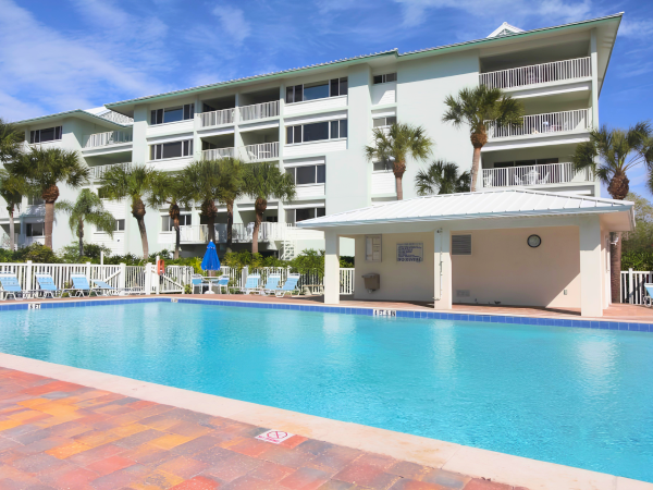 A sunny hotel with a large blue swimming pool, palm trees, and a multi-story white building in the background, sunny day and clear sky.