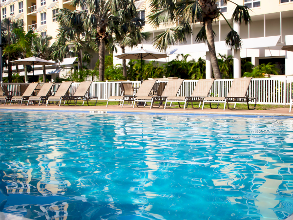 A resort pool with clear blue water, lounge chairs lined up by a white fence, and palm trees in the background, sunny day.