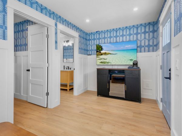 A bright hotel-style room with blue patterned wallpaper, white wainscoting, a wooden floor, a black TV stand with a flat-screen, and a dresser, plus a doorway leading to another room.