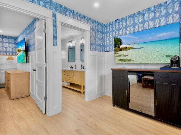 A hotel suite living area with bright blue patterned walls, a flat-screen TV, a dark cabinet, and doorways leading to a bright bathroom and bedroom.