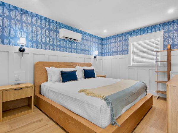 A modern hotel-style bedroom with a wooden bed, blue patterned wallpaper, white wainscoting, and light wood furniture.