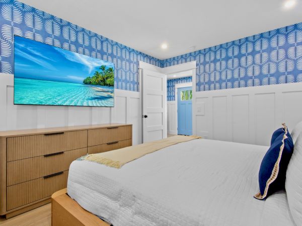 A bright bedroom with a mounted TV, blue patterned wallpaper, white wainscoting, a wooden dresser, and a neatly made bed. (140 chars)