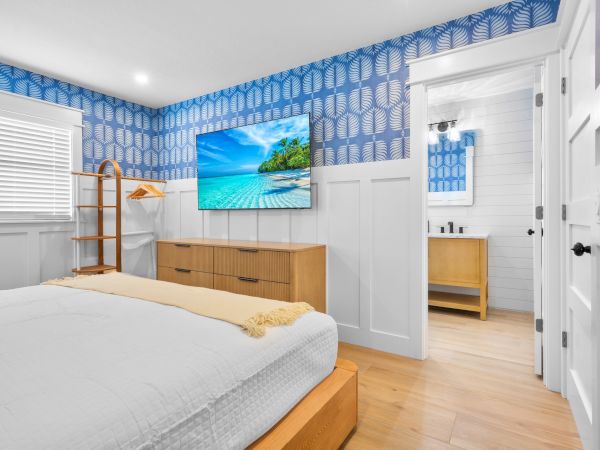 A bright bedroom with blue patterned wallpaper, white panel accents, a wooden bed frame, dresser, wall-mounted TV, and a small dresser mirrored in an attached space.