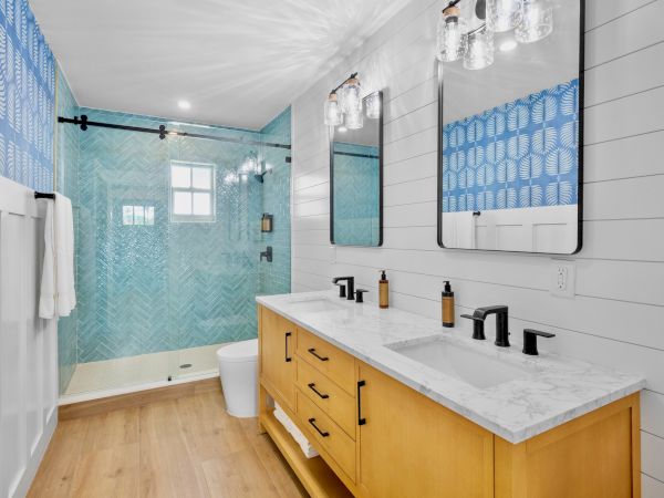 A bright bathroom with a double vanity, light wood cabinet, white countertop, two mirrors, blue patterned shower area, glass light fixtures, and wooden floor.