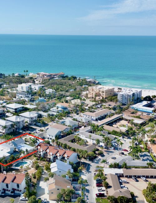 Aerial view of a coastal town with houses, palm trees, and grid-like streets next to a bright blue ocean and shoreline.
